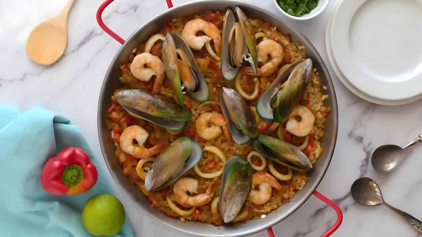 A seafood paella in a red-handled pan, topped with shrimp, mussels, and squid rings. Beside it are a red pepper, lime, wooden spoon, empty plates, and a small bowl of herbs.