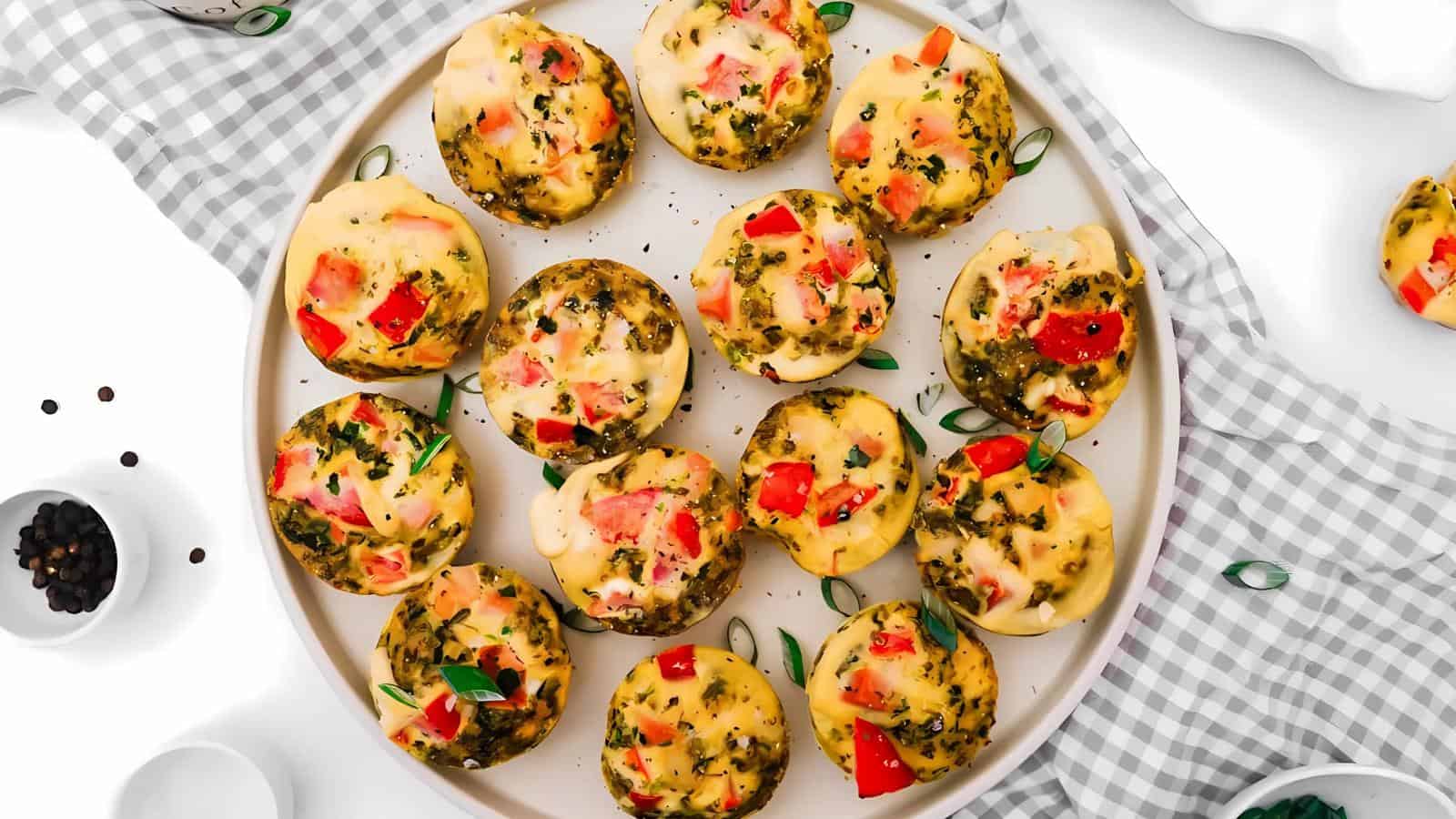 A plate of small, round, baked egg muffins with melted cheese, diced red bell peppers, and herbs, displayed on a checkered cloth with a few peppercorns nearby.