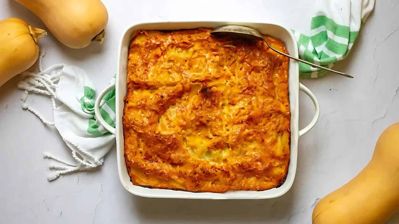 A baked dish in a square white casserole, topped with melted cheese, surrounded by butternut squash and a green-striped kitchen towel.