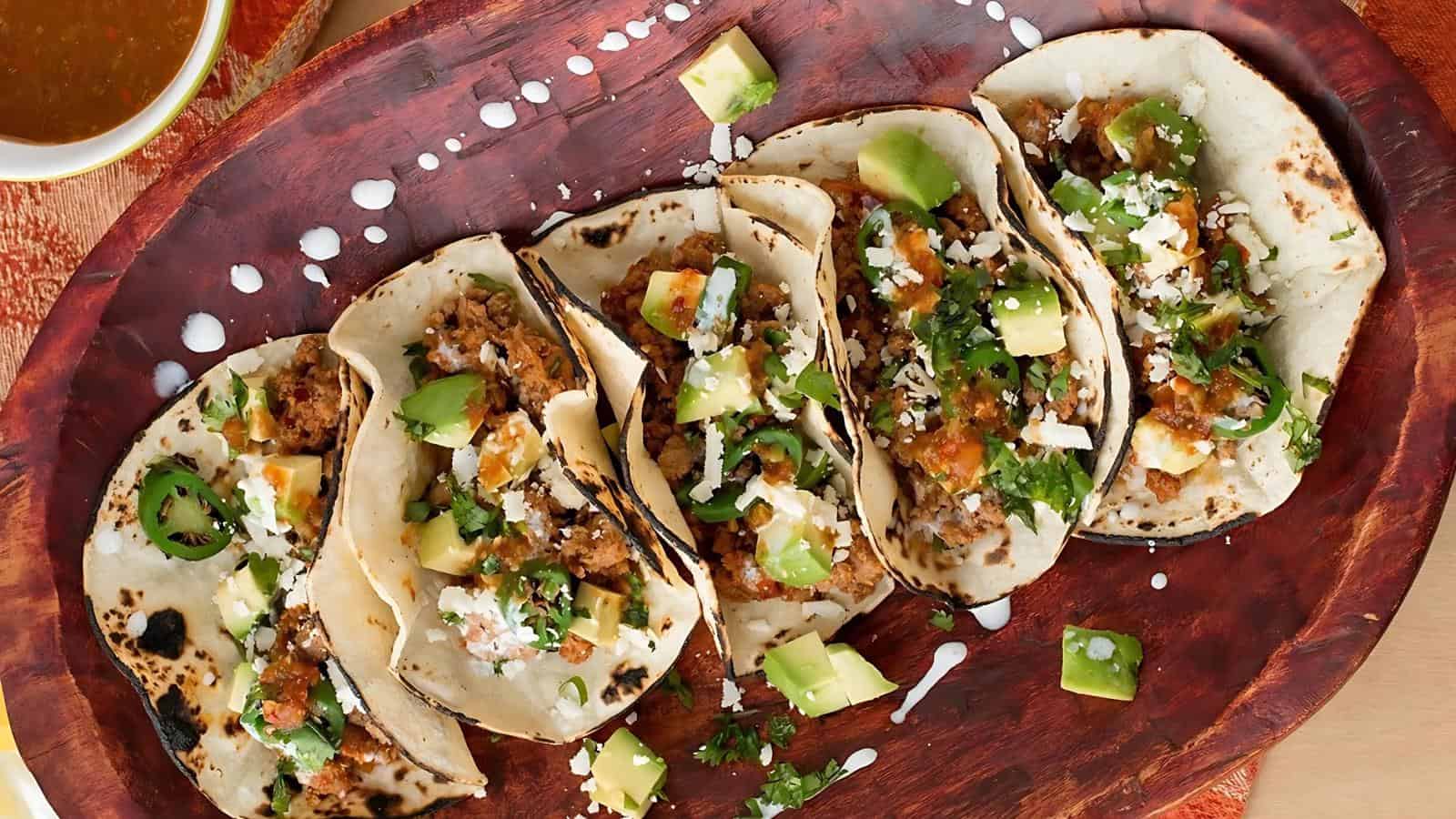 Six tacos filled with meat, avocado, cilantro, cheese, and sauce on a wooden platter.