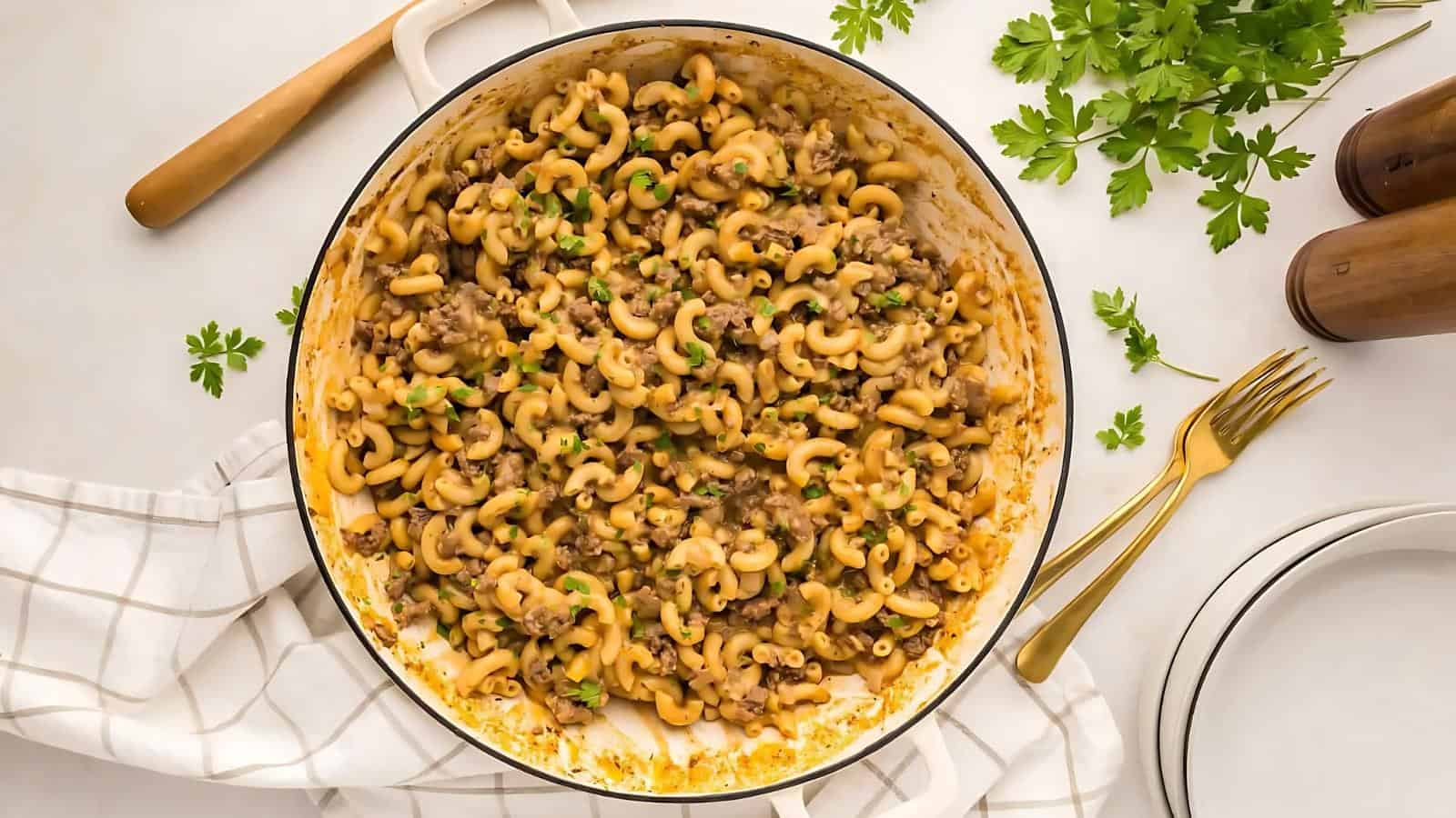 A pot of cooked macaroni mixed with ground meat and garnished with chopped herbs, placed on a checked cloth. A fork, knife, plates, and parsley surround the pot.
