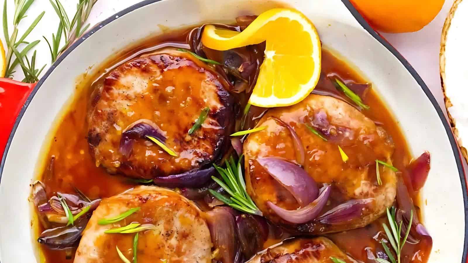 Pork chops in a pan with sliced onions, rosemary, and a glossy sauce, garnished with a slice of orange.