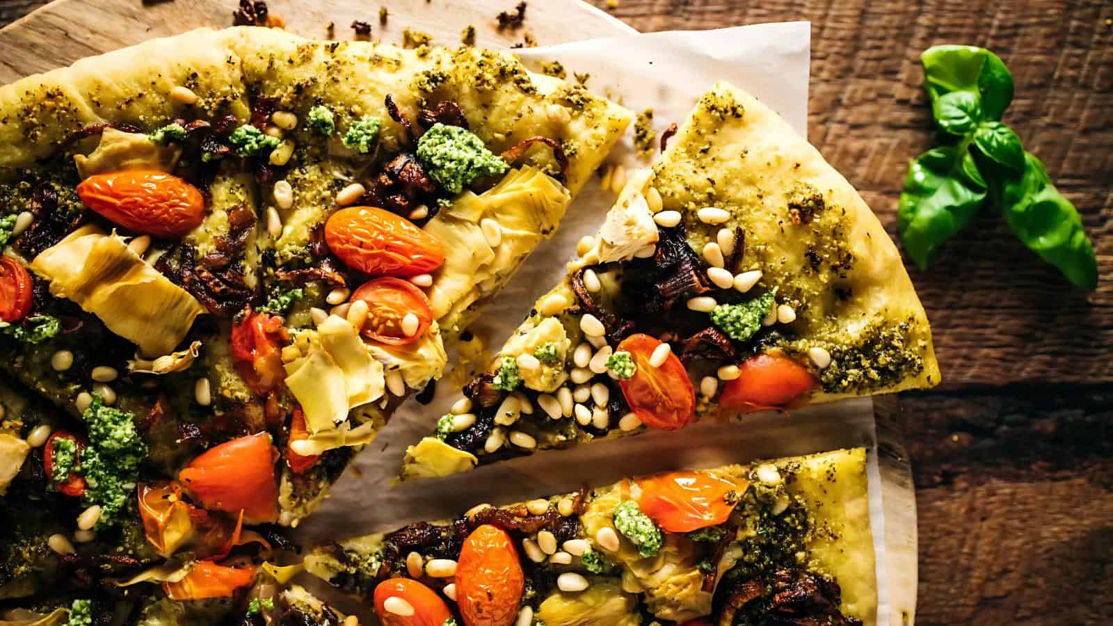 Close-up of a pesto pizza topped with cherry tomatoes, artichokes, mushrooms, pine nuts, and dollops of green sauce, on a wooden surface beside a sprig of fresh basil.