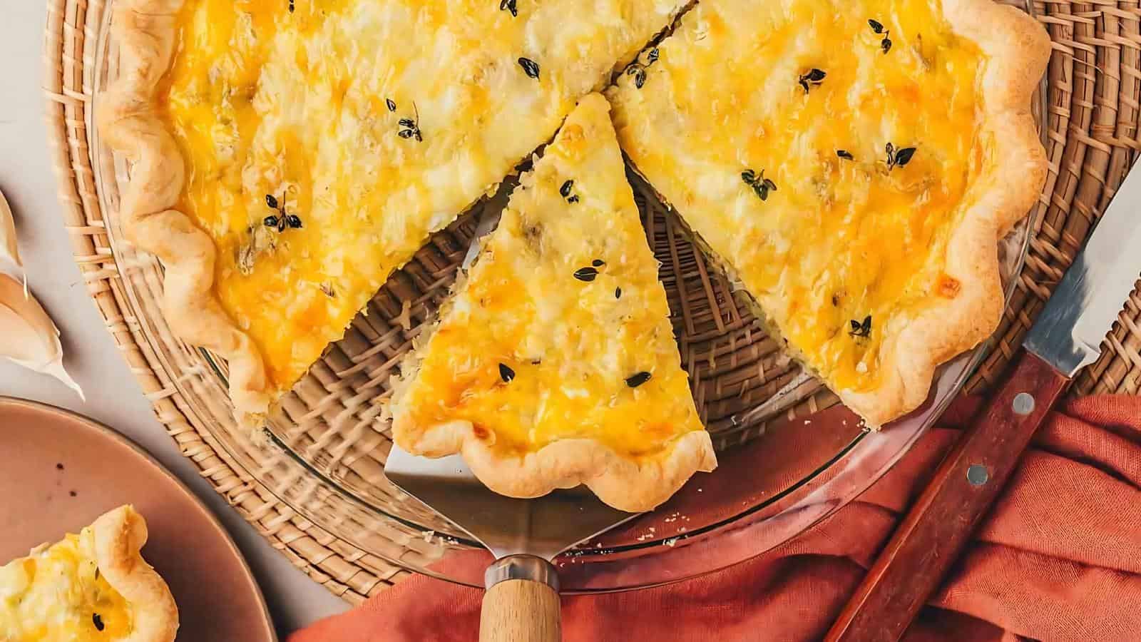 A sliced cheese quiche in a glass pie dish on a woven mat with a serving spatula. A red napkin and a plate with another slice are visible nearby.