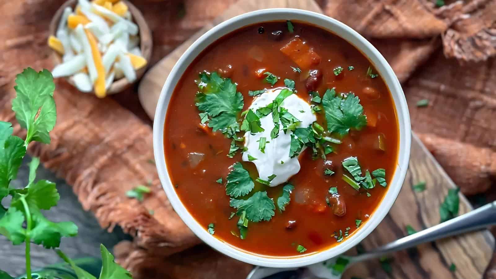 A bowl of chili topped with sour cream and cilantro on a rustic cloth background.
