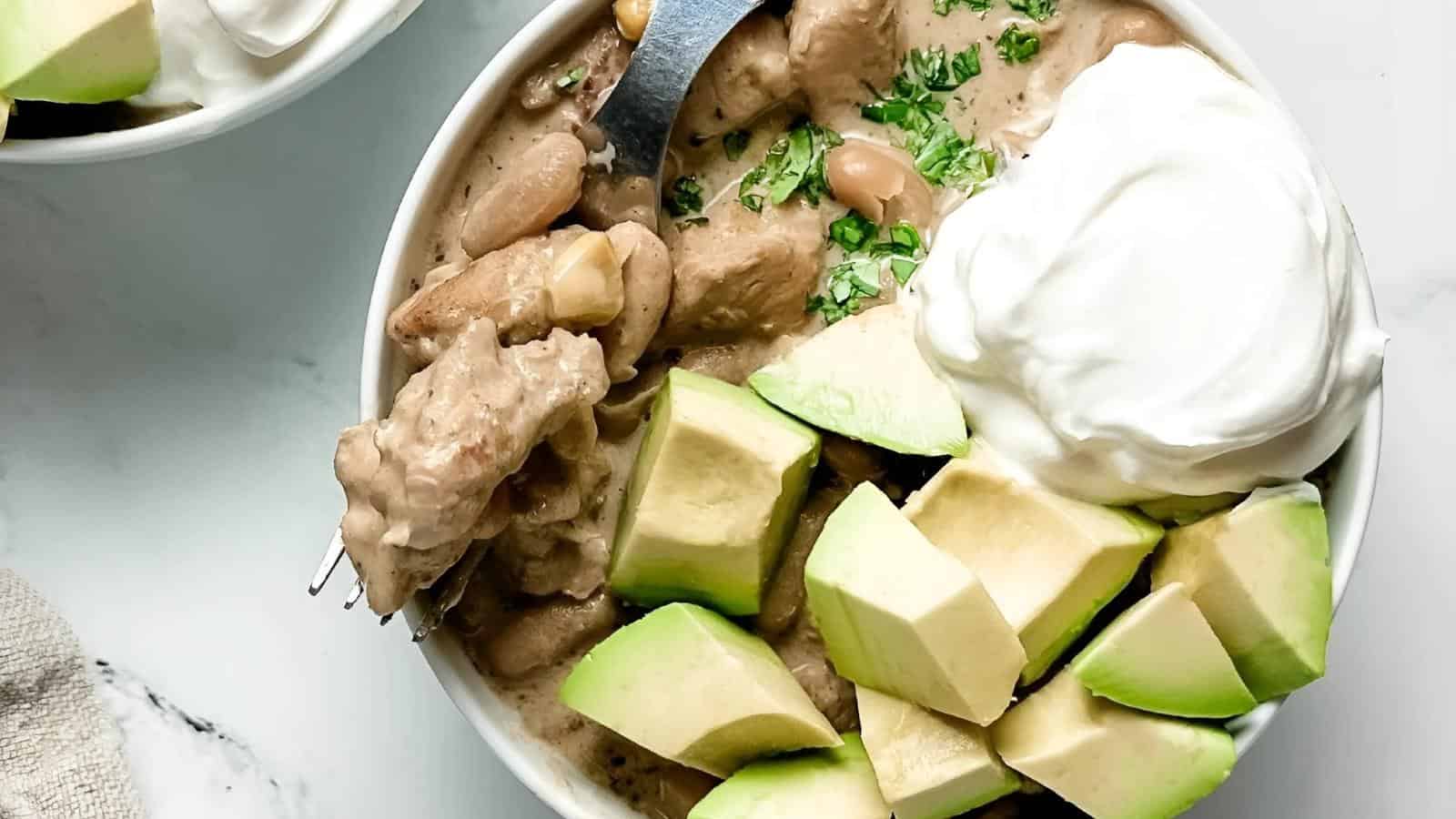 A bowl of bean stew topped with avocado chunks, sour cream, and chopped herbs, with a fork resting inside.