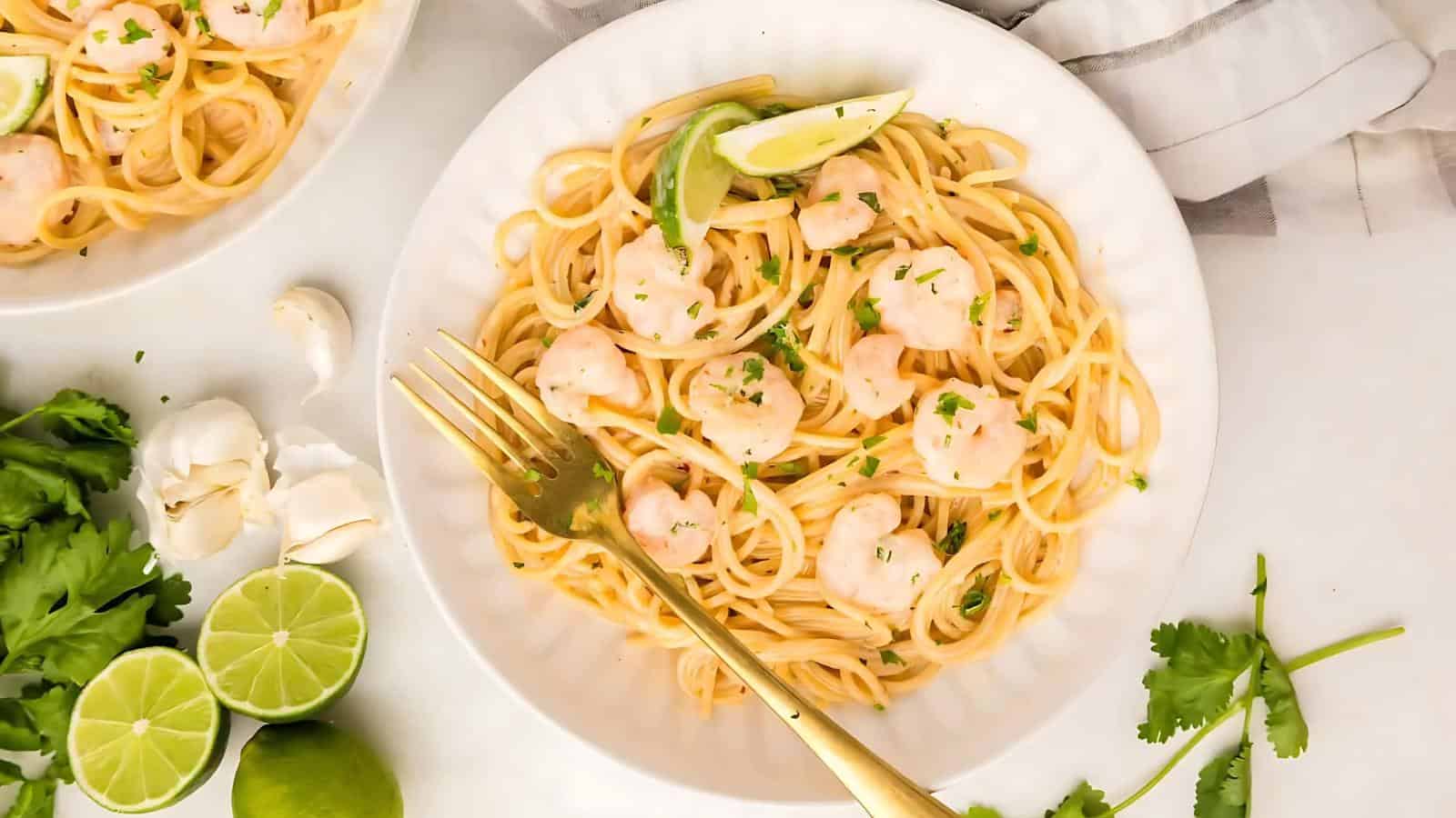 A plate of shrimp pasta garnished with lime wedges and parsley, accompanied by garlic cloves and fresh cilantro on the side.
