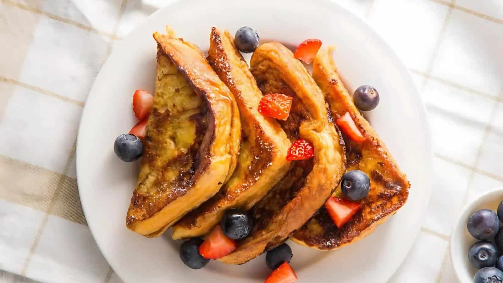 Four slices of French toast topped with blueberries and strawberry pieces on a white plate.