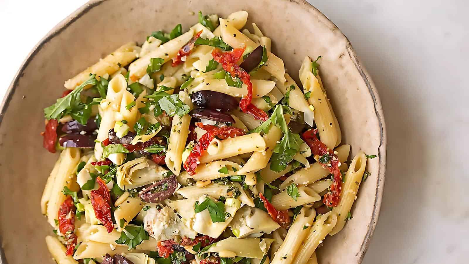 A bowl of penne pasta salad with olives, sun-dried tomatoes, artichokes, and parsley on a light-colored surface.