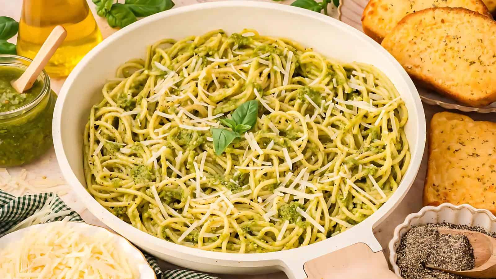 A pan of spaghetti topped with green pesto and grated cheese, surrounded by garlic bread, a jar of pesto, olive oil, and a bowl of grated cheese.