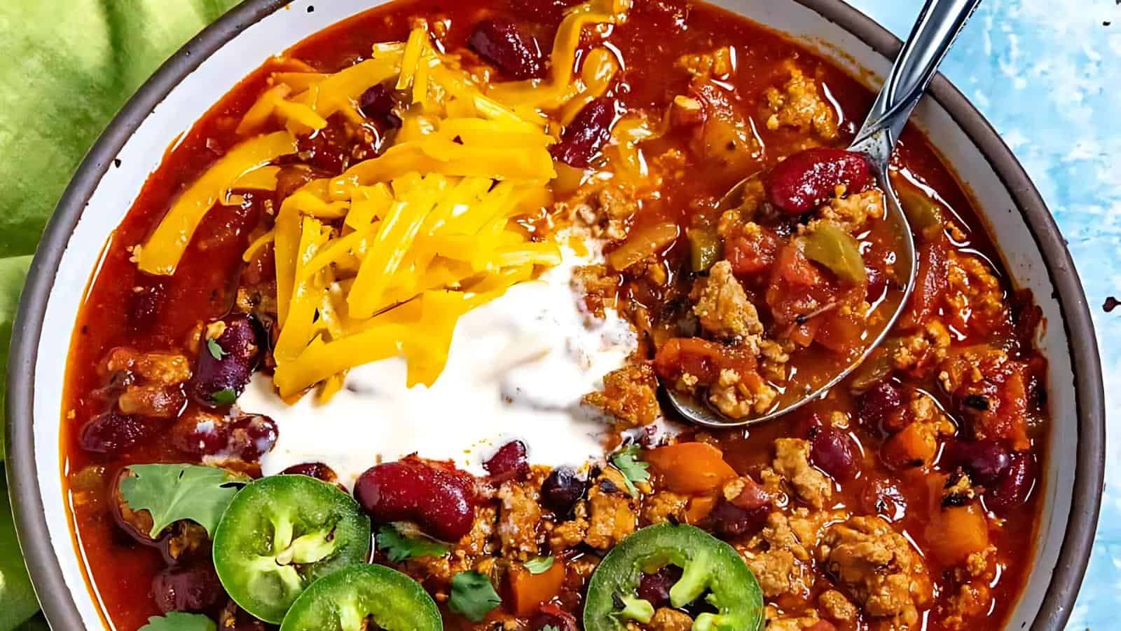 A bowl of chili with ground meat, red beans, and tomato sauce, topped with shredded cheddar cheese, sour cream, and sliced jalapeños, with a spoon on the side.