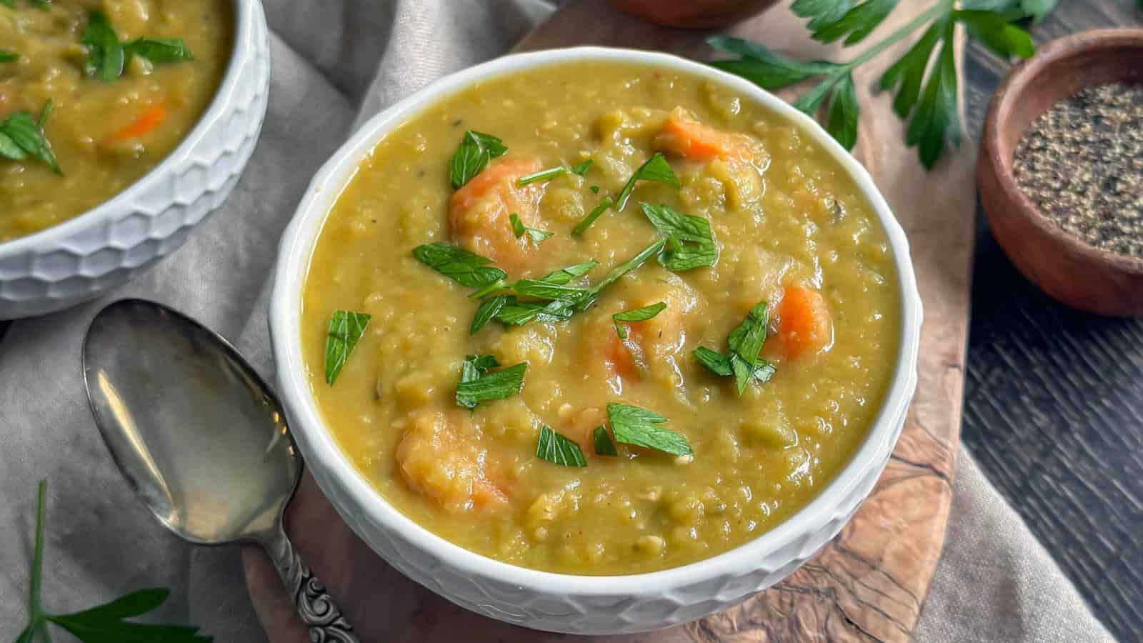 A bowl of split pea soup with carrot pieces, garnished with fresh parsley, on a wooden board next to a spoon and a small bowl of pepper.