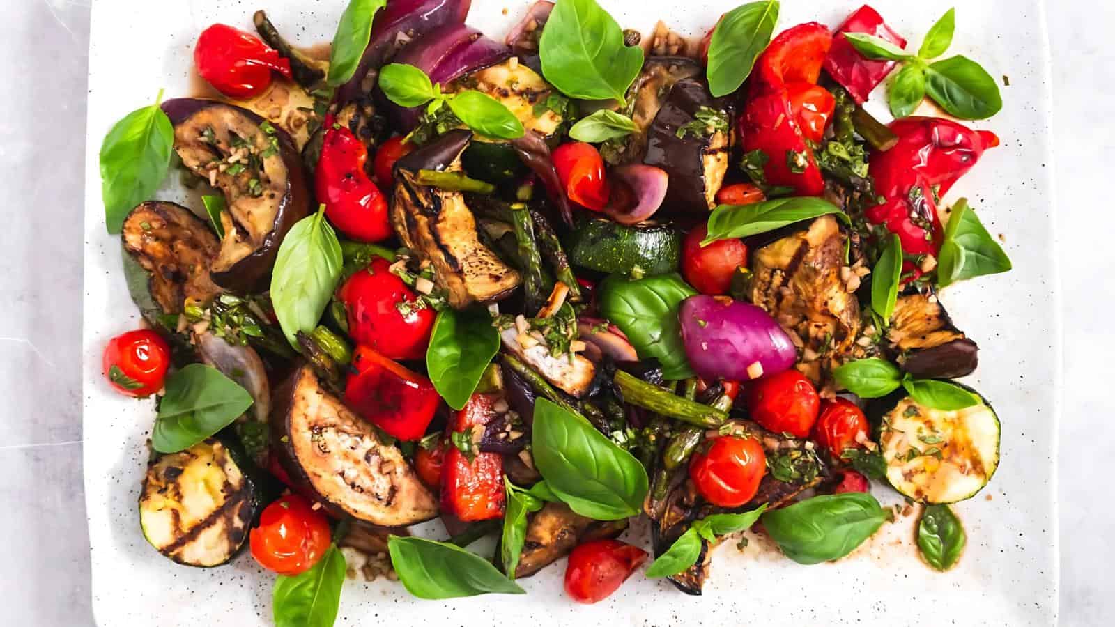 A colorful assortment of roasted vegetables, including eggplant, zucchini, asparagus, and cherry tomatoes, garnished with fresh basil leaves on a white plate.