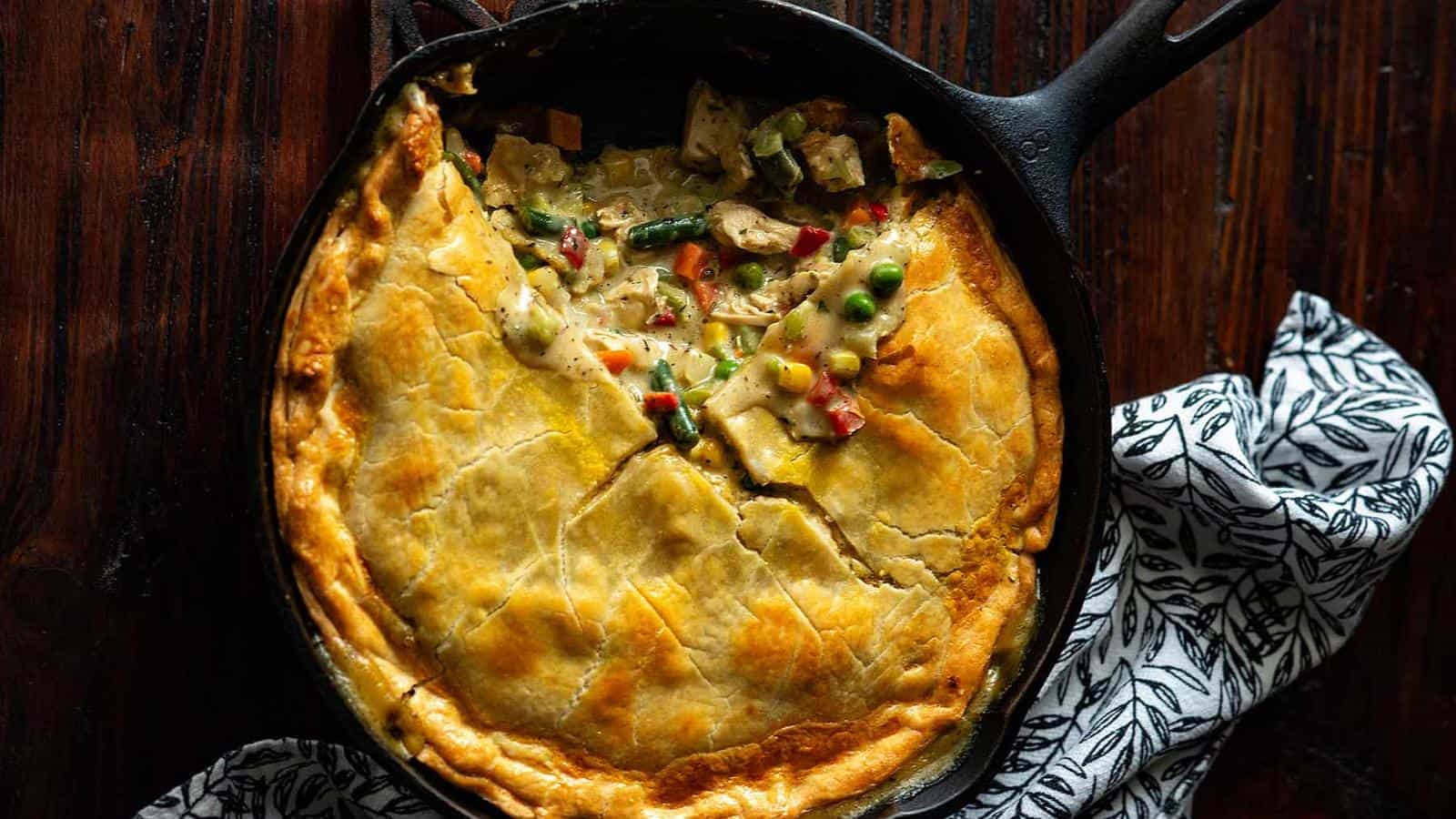 A pot pie with a golden-brown crust and visible mixed vegetable filling, in a black skillet on a wooden surface, partially wrapped with a patterned cloth.