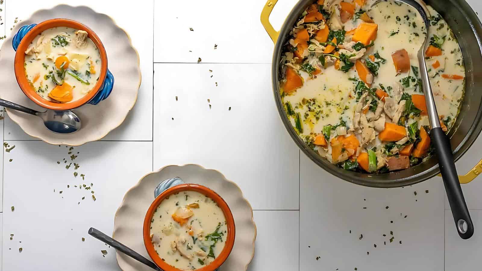 A pot of creamy soup with chicken, sweet potatoes, and greens. Two bowls of the soup are served on plates with spoons on the side. Sprinkled herbs are scattered on the white surface.