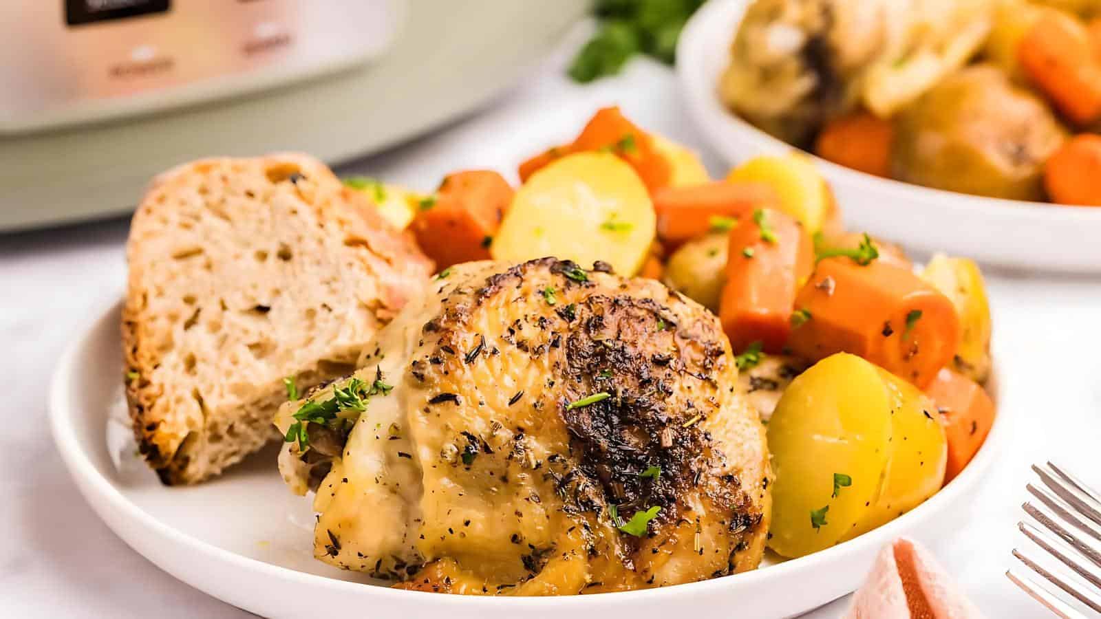 Plate of roasted chicken with carrots and potatoes, garnished with herbs, alongside a slice of bread.