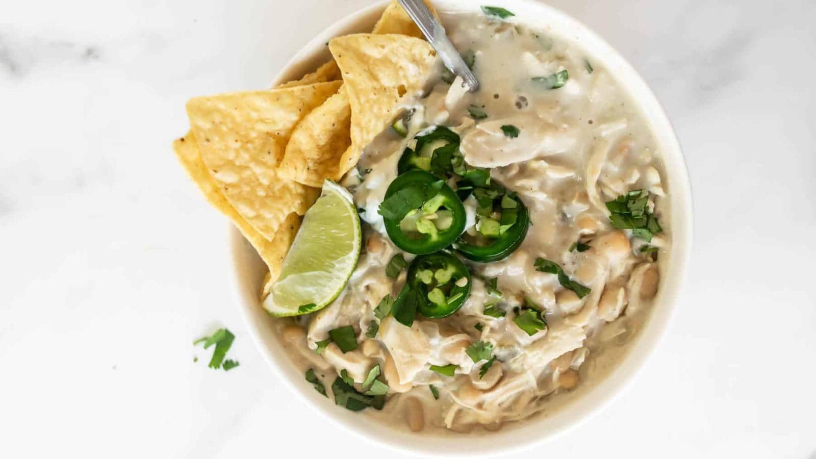 A bowl of white chicken chili topped with sliced jalapeños, tortilla chips, and a lime wedge.