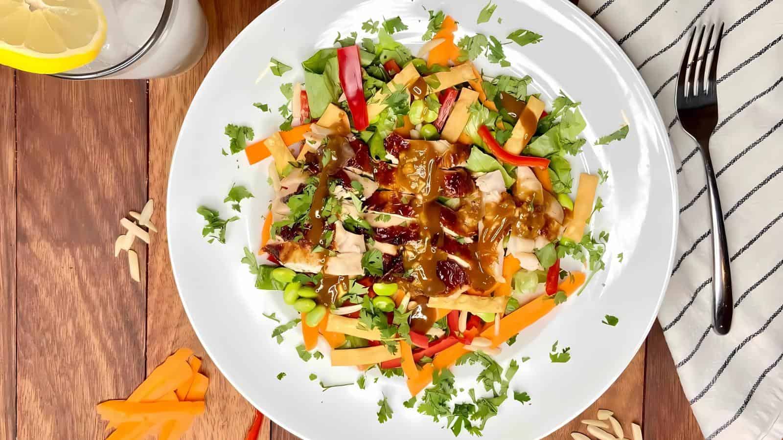 A colorful salad on a white plate featuring mixed greens, red and orange bell peppers, shredded carrots, and nuts, topped with dressing. A drink and napkin with fork are in the background.