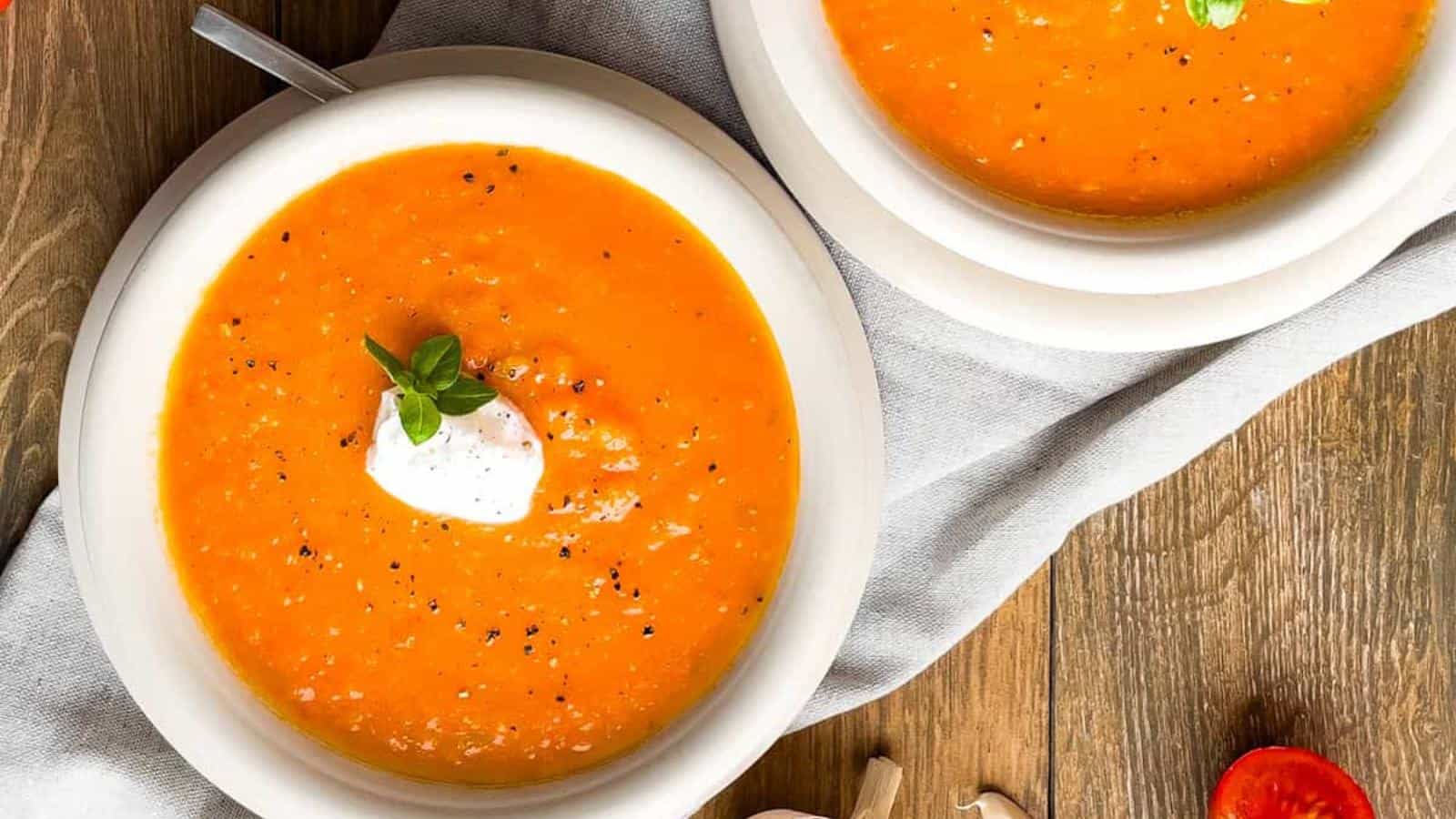 Two bowls of tomato soup topped with sour cream and herbs on a wooden table. Garlic cloves and a sliced tomato are nearby.