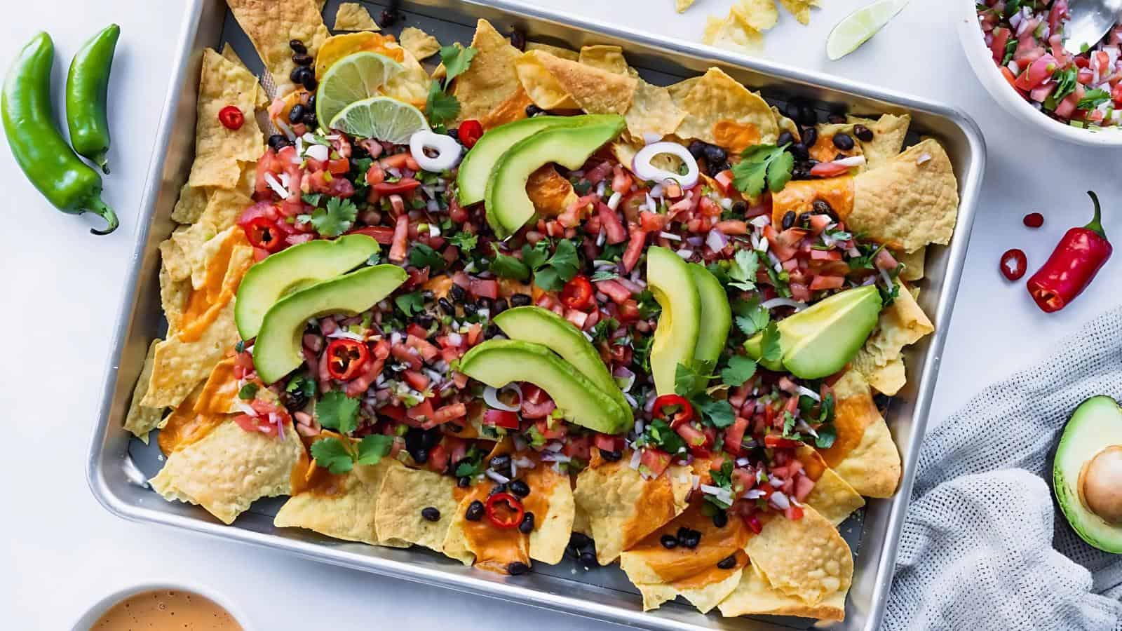 A tray of nachos topped with sliced avocado, diced tomatoes, cilantro, jalapeños, lime wedges, and onion, surrounded by ingredients like chili peppers and an avocado on a white surface.