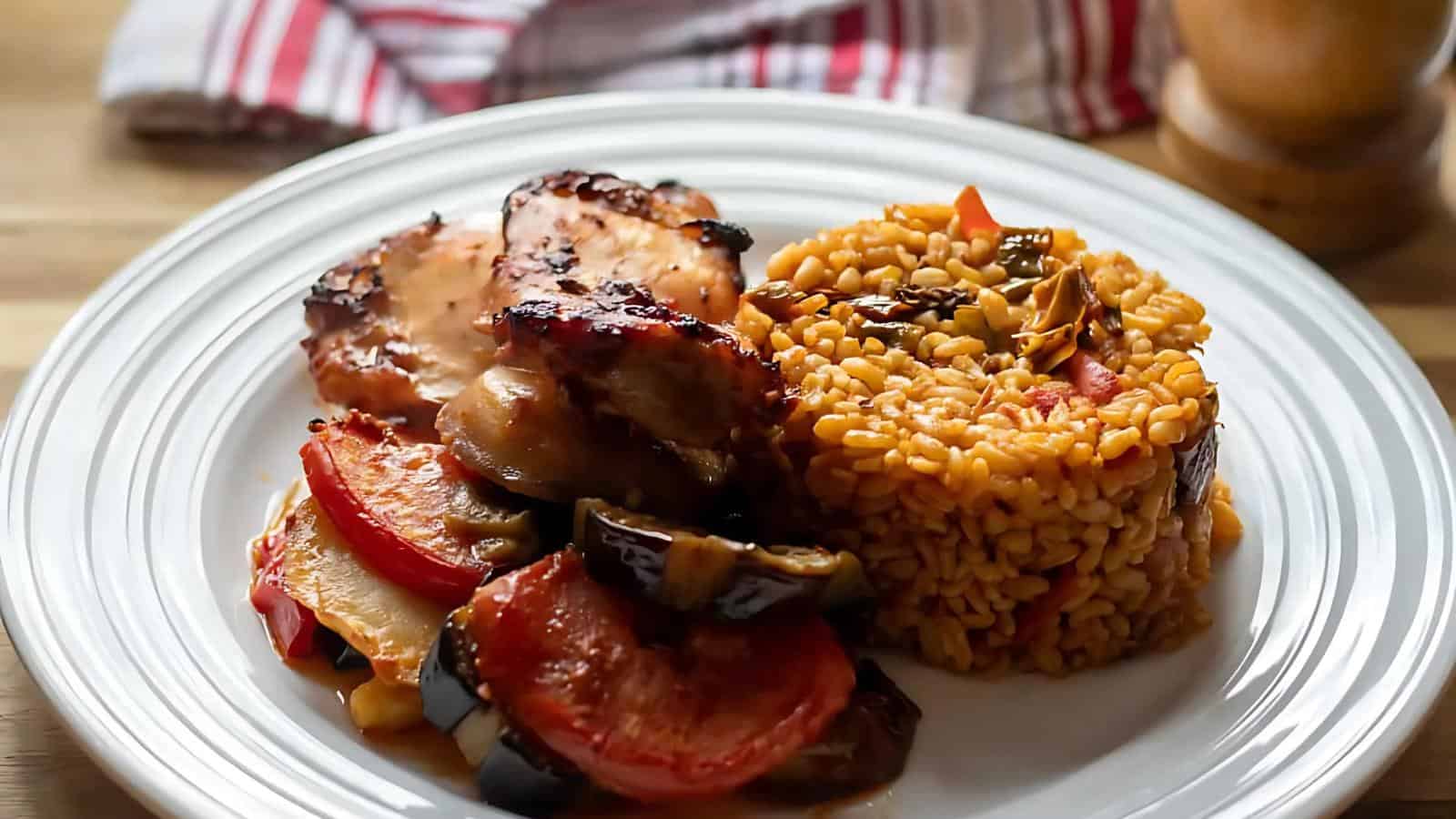 A white plate with cooked rice, sliced grilled vegetables, and roasted chicken, placed on a wooden table. A striped cloth is visible in the background.