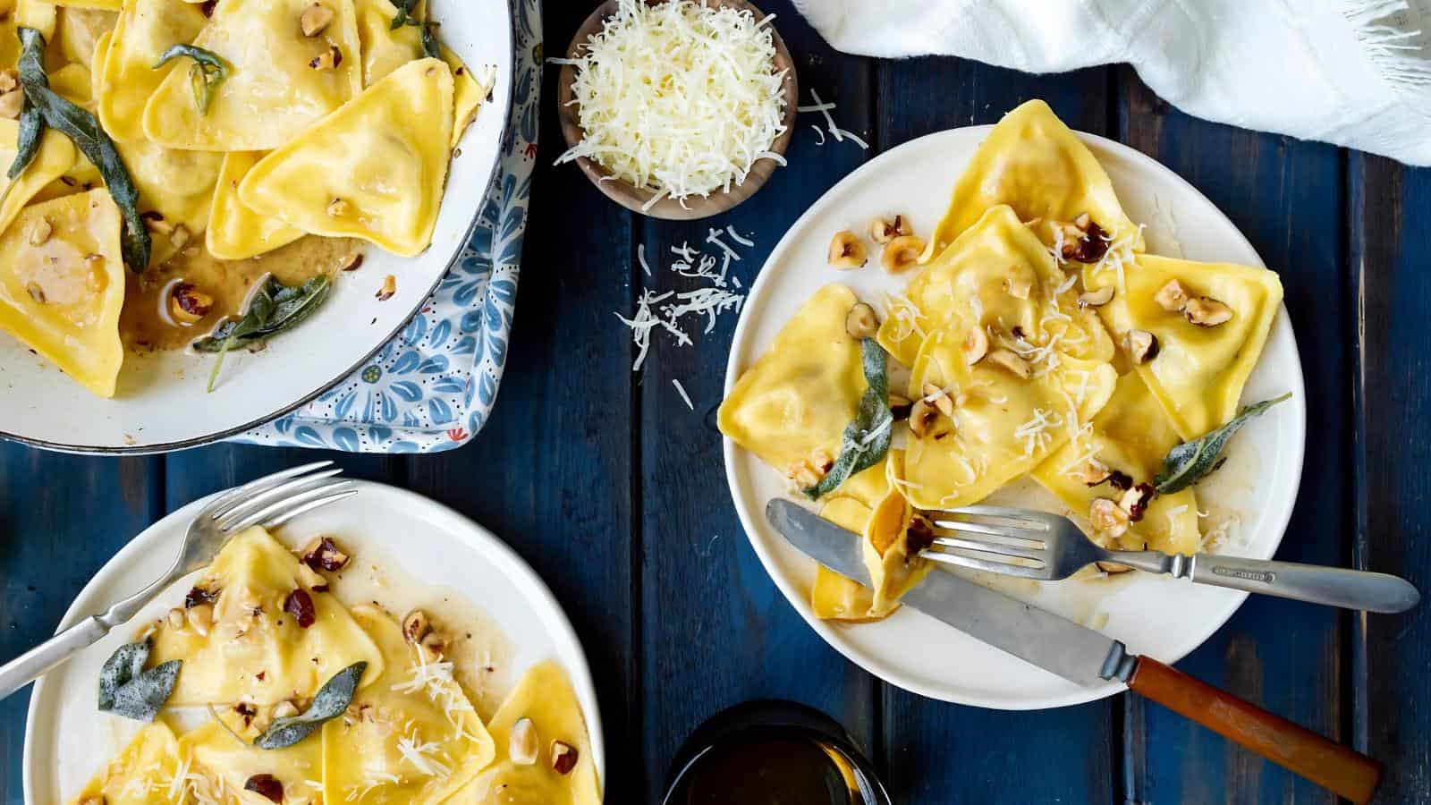 Two plates of ravioli with brown butter sauce, topped with sage and nuts, next to a bowl of grated cheese on a dark wooden table.