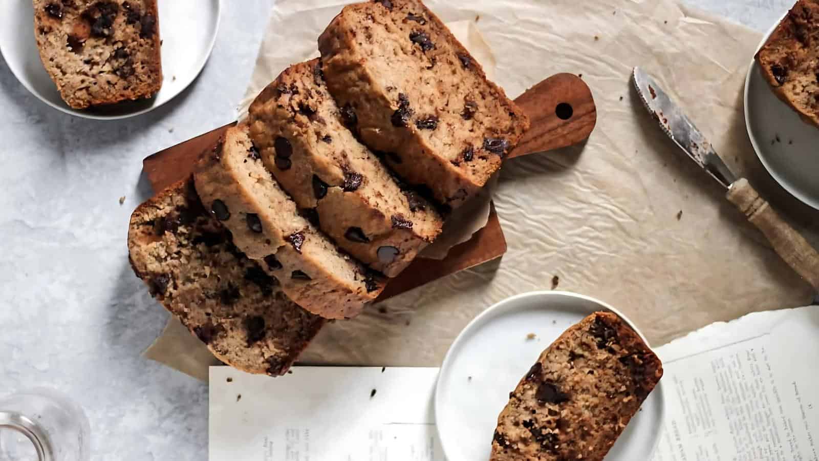 Slices of chocolate chip bread on a wooden board, surrounded by plates, a knife, and paper on a light surface.