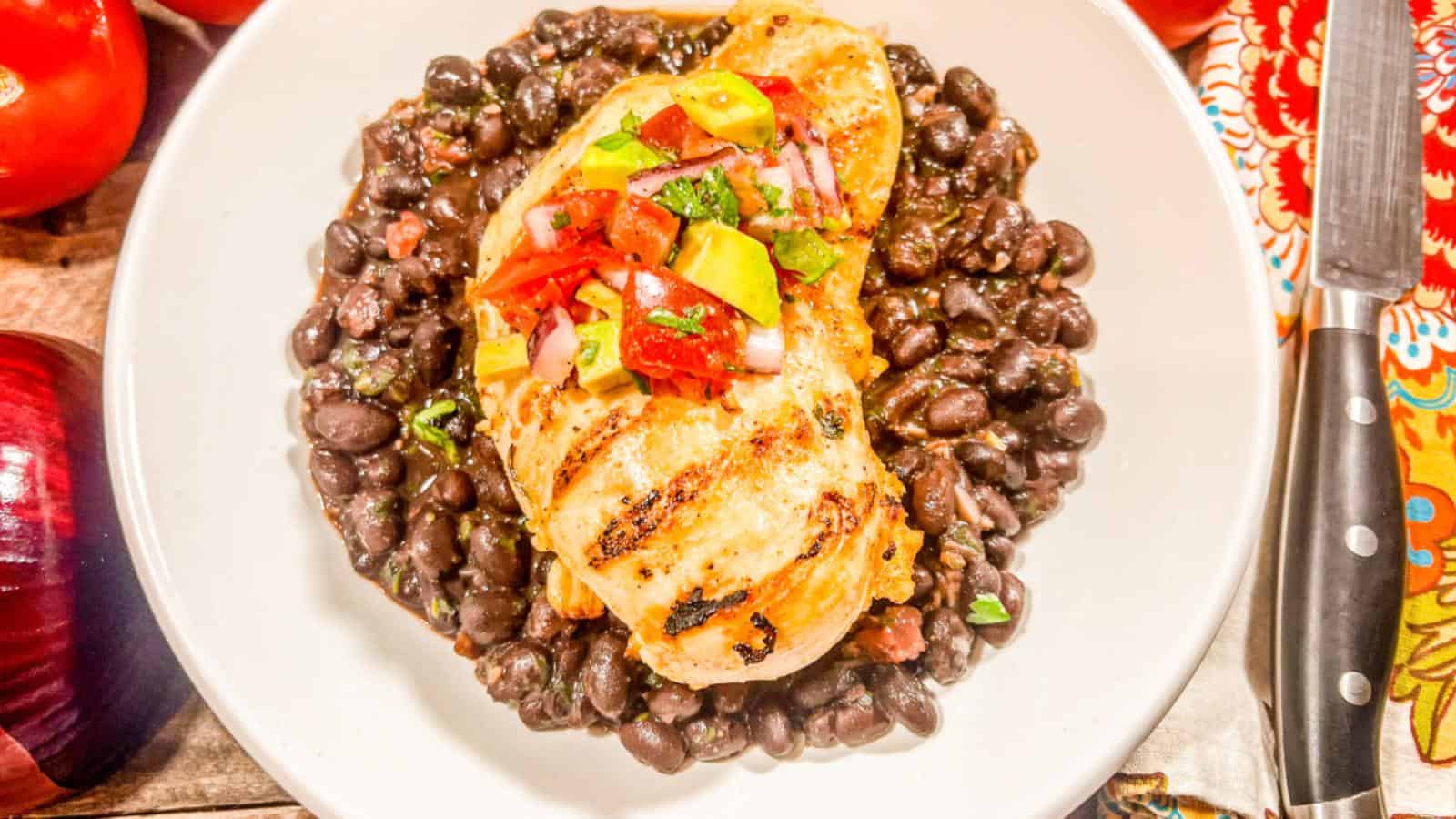 Grilled chicken breast topped with salsa on a bed of black beans, served in a white bowl. A knife is placed beside the bowl.