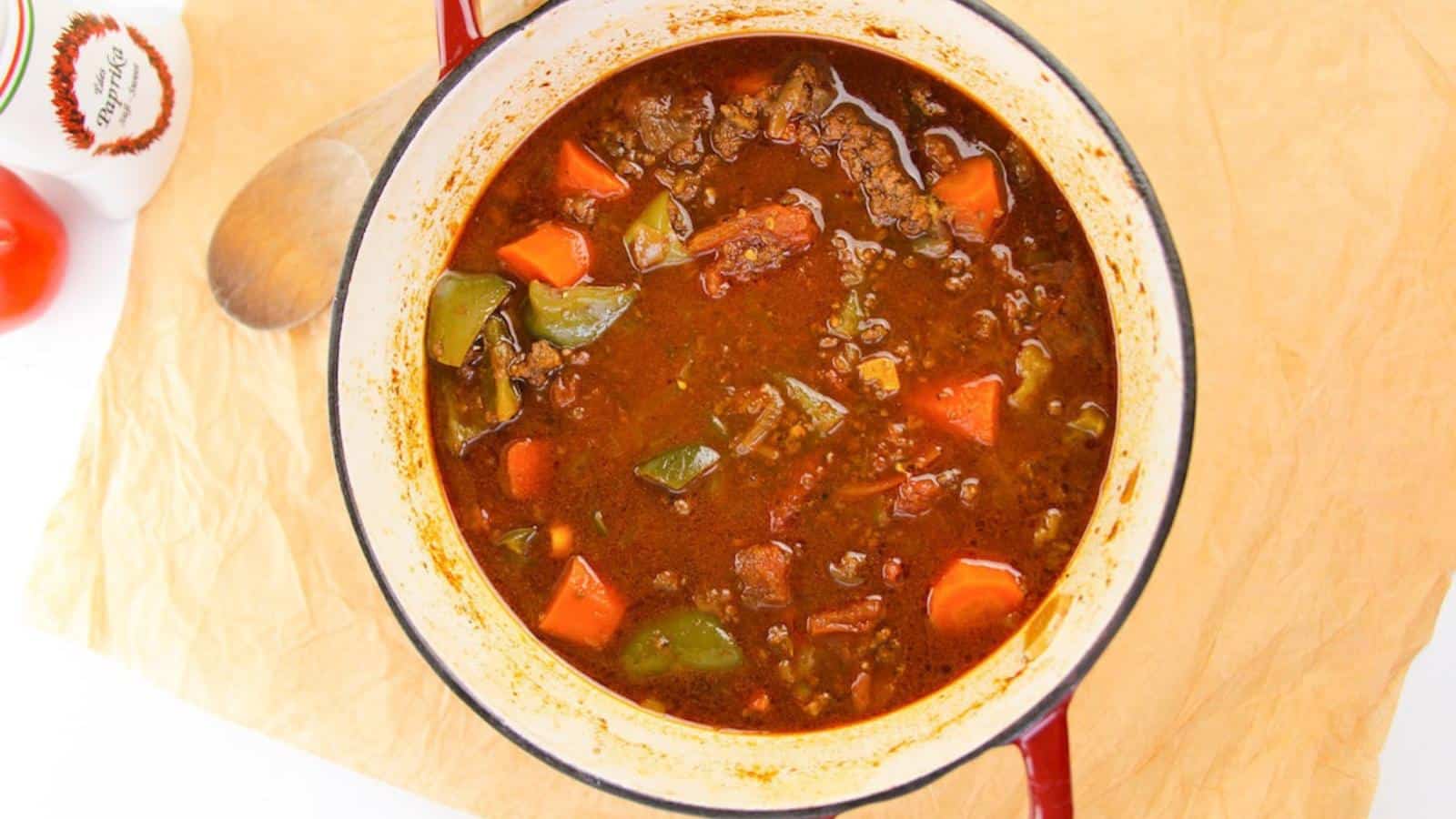 A pot of beef stew with chunks of meat, carrots, green peppers, and a rich broth on a wooden surface.