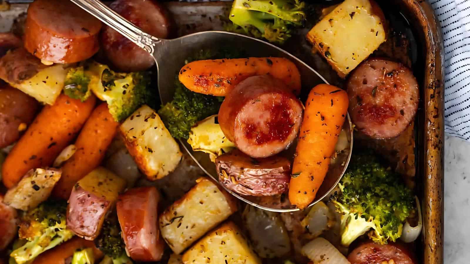 Baked vegetables and sausage chunks on a tray, featuring carrots, potatoes, broccoli, and herbs, with a serving spoon lifting a portion.