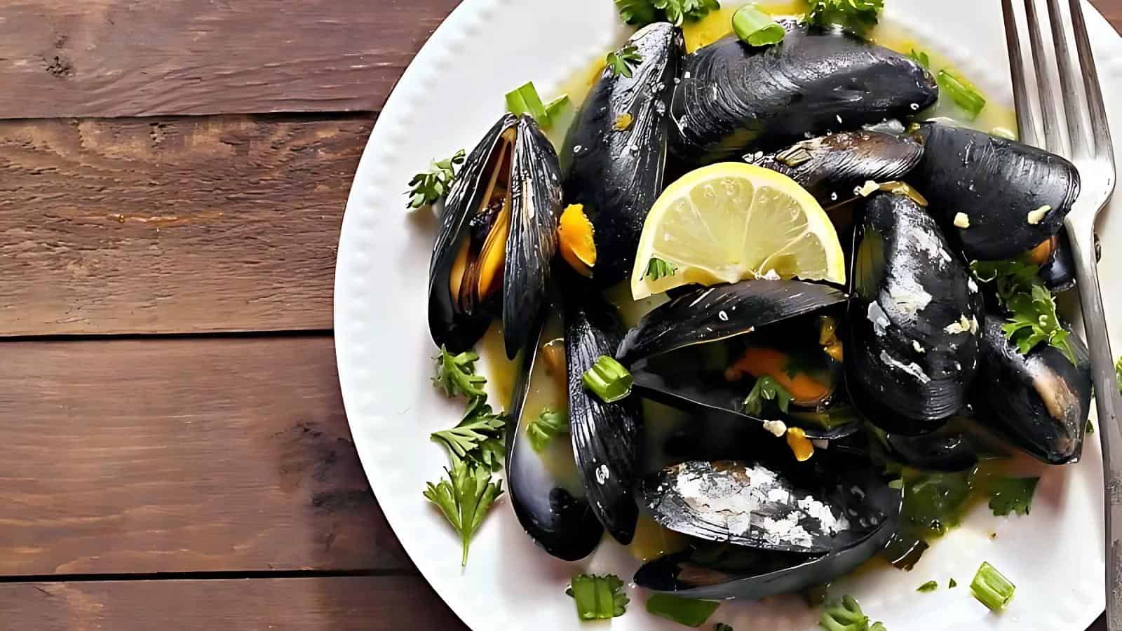 A white plate with cooked mussels garnished with lemon slices, chopped green onions, and parsley is placed on a wooden surface. A fork is on the right side of the plate.
