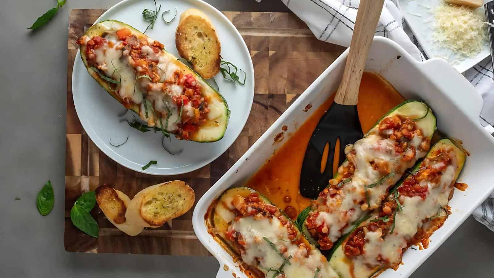 Zucchini boats filled with vegetables and topped with cheese, served in a white baking dish, with a plate on the side. Toasted bread slices accompany the meal on a wooden board.