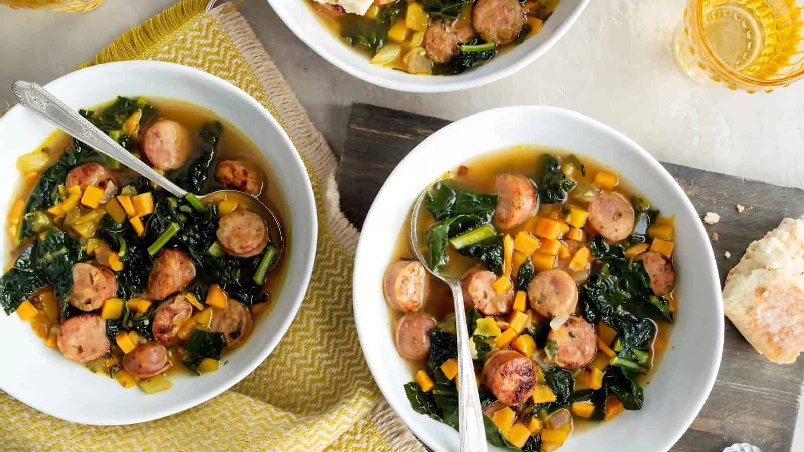 Three bowls of sausage and vegetable soup with kale, yellow peppers, and broth on a table with a yellow cloth. Spoons are in the bowls, and there is bread beside one bowl.