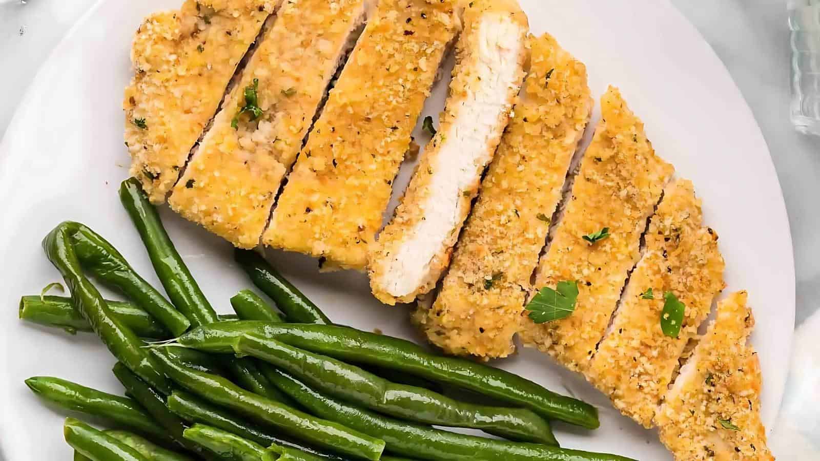 Sliced breaded chicken breast with herbs next to cooked green beans on a white plate.