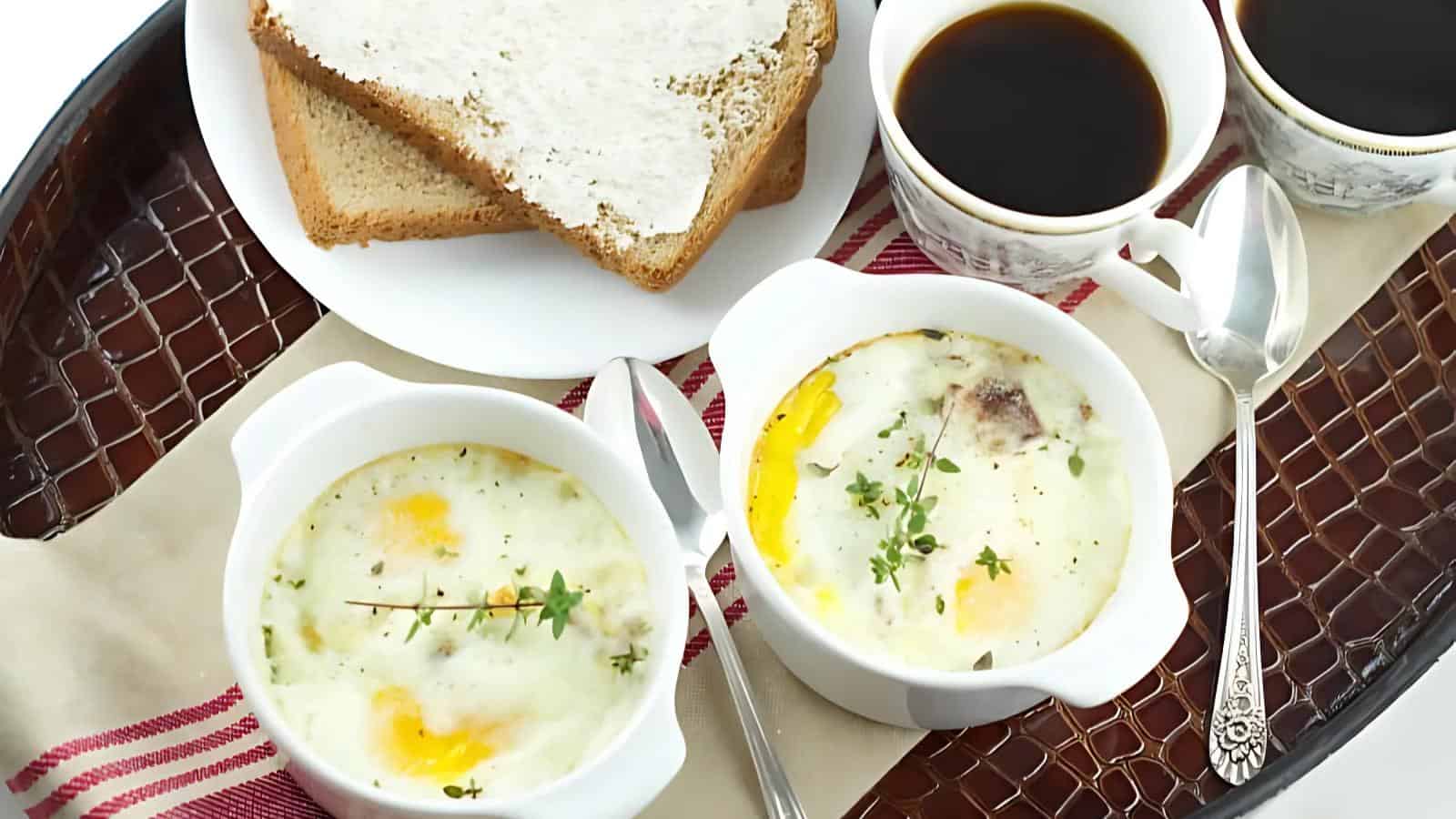 Two bowls of baked eggs topped with herbs, two cups of black coffee, and buttered toast on a tray.