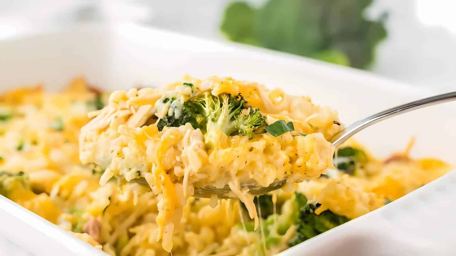 A spoon lifts a portion of chicken and broccoli casserole topped with melted cheese from a white baking dish.