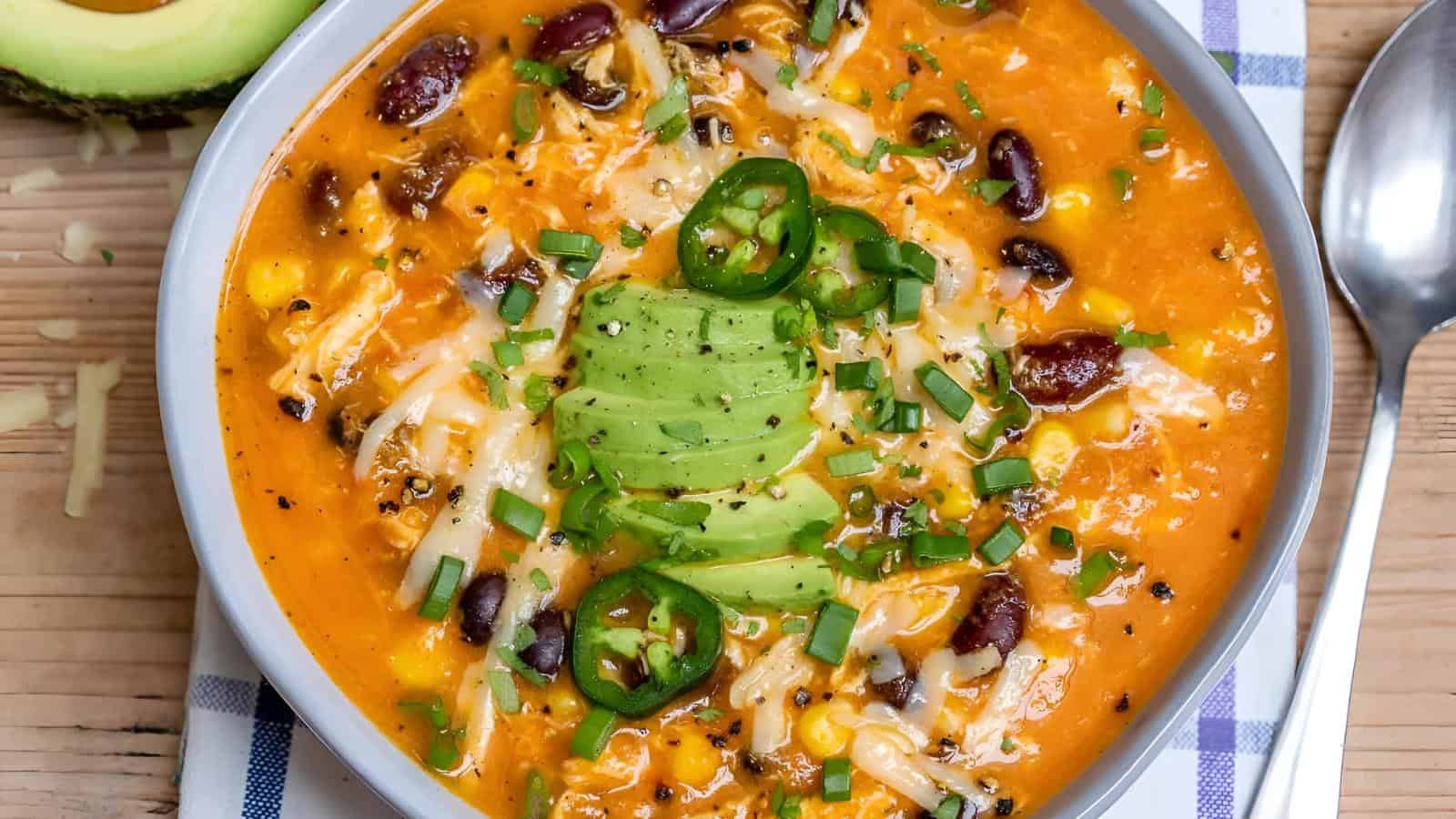 A bowl of chili soup topped with sliced avocado, jalapeños, grated cheese, and chopped chives. A spoon is placed beside the bowl on a wooden table.
