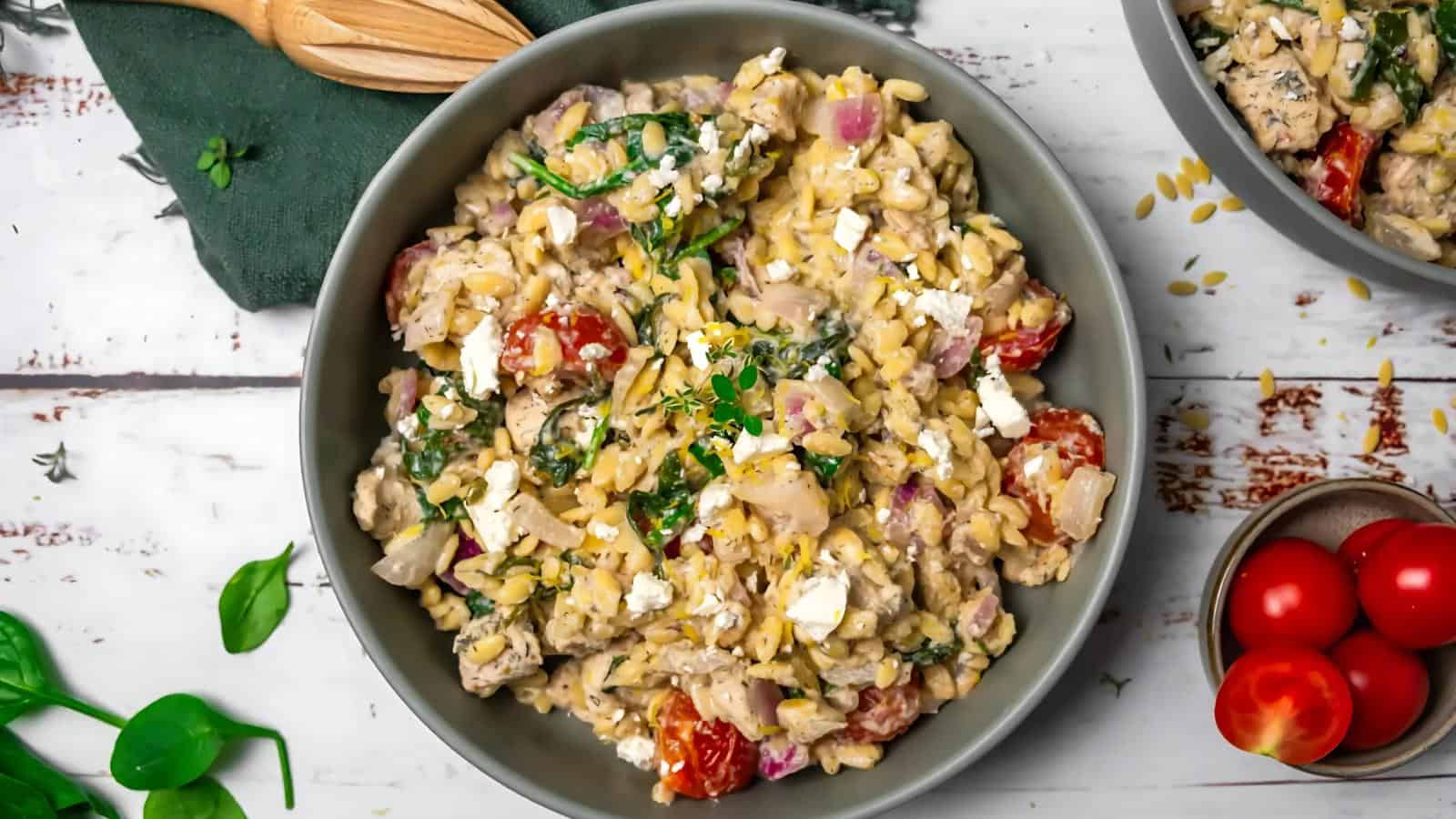 A bowl of creamy orzo pasta with spinach, cherry tomatoes, red onions, cubes of chicken, and crumbled feta cheese, garnished with herbs.