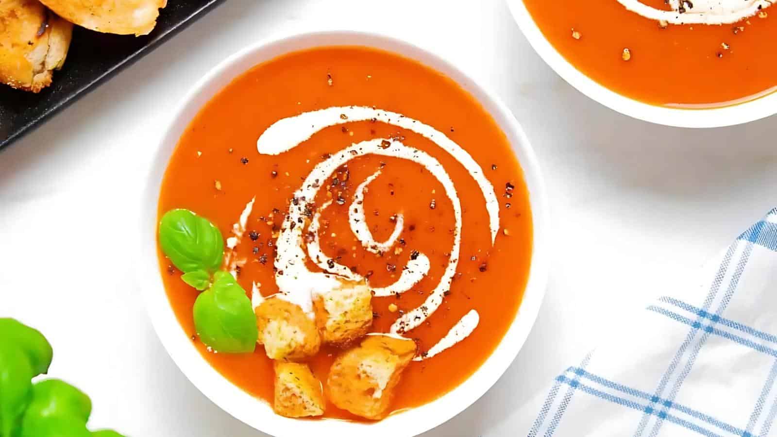 A bowl of tomato soup garnished with a swirl of cream, black pepper, croutons, and a basil leaf, placed on a white surface near a blue-and-white checkered cloth.