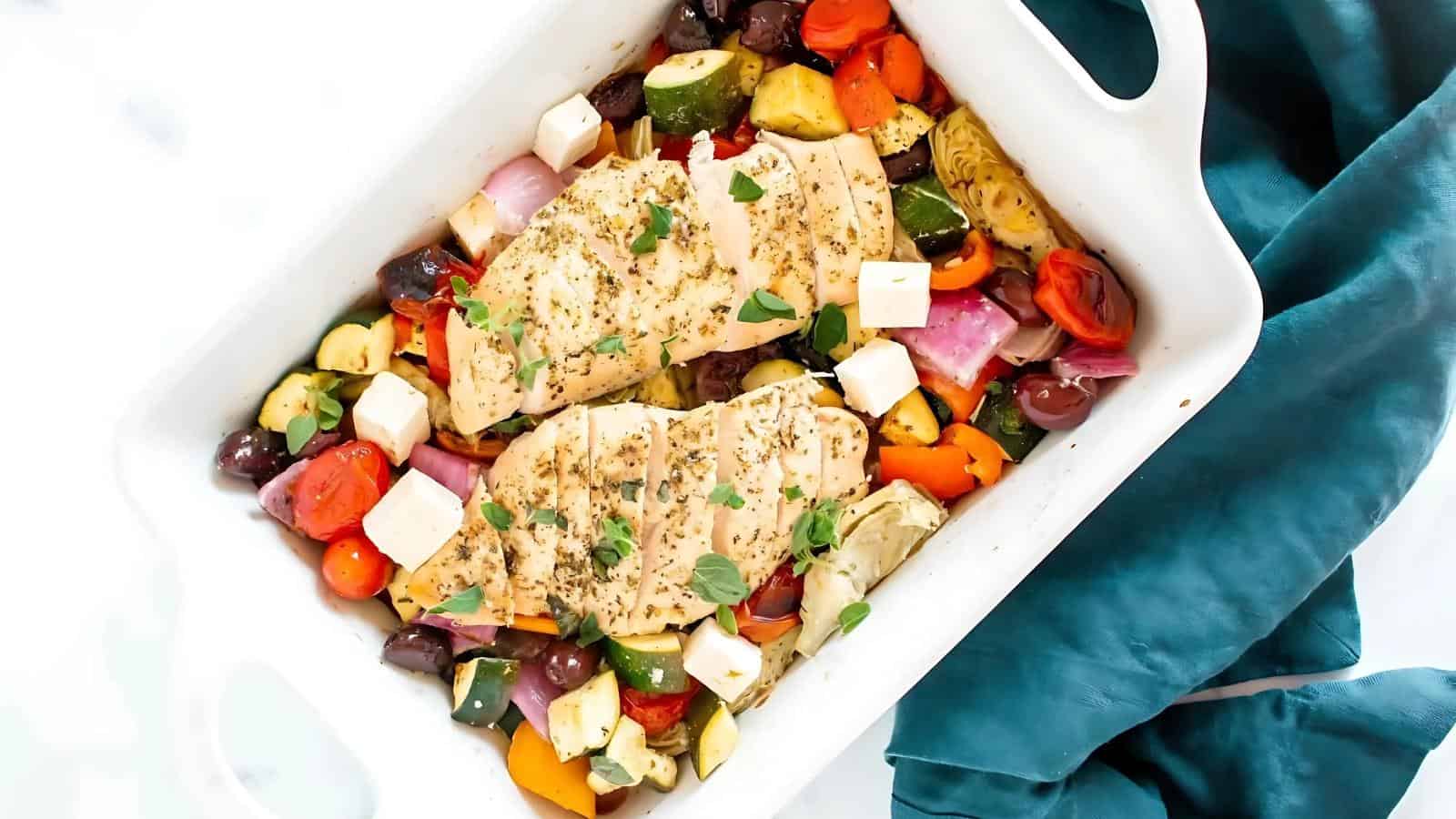 A white dish with baked chicken breasts, chopped vegetables, olives, and cubes of cheese, garnished with herbs. A blue cloth is partially visible on the side.