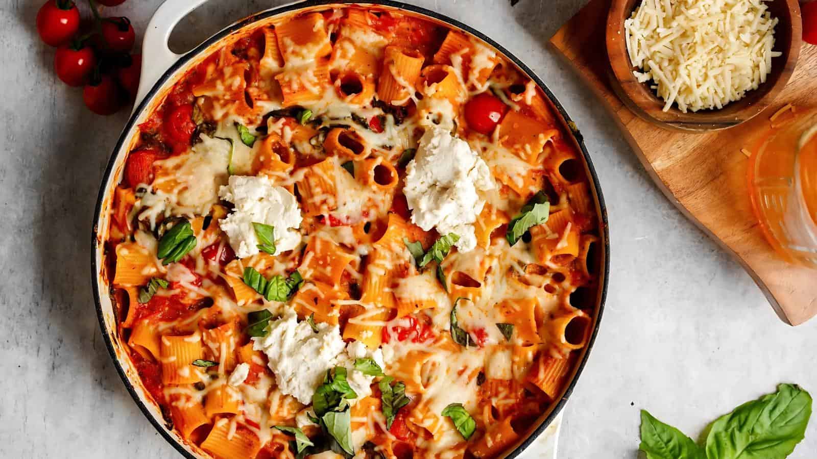 A baked pasta dish with rigatoni, melted cheese, tomatoes, and basil in a pan. A bowl of grated cheese and cherry tomatoes are nearby.