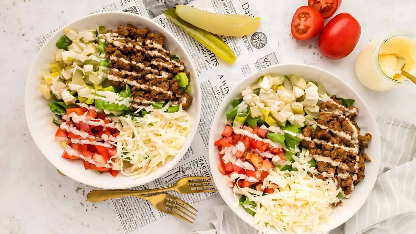 Two bowls of salad with shredded cheese, diced tomatoes, avocado, crumbled meat, and dressing drizzled on top, placed on a newspaper with forks beside them.