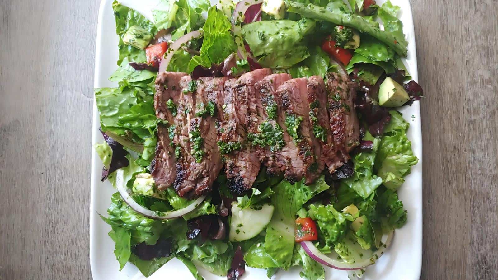 Sliced grilled steak on a bed of mixed leafy greens with avocado, cucumber, red onion, and red bell pepper, garnished with herbs.