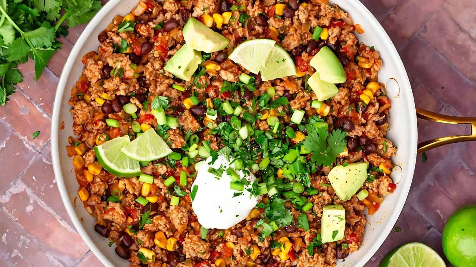 A bowl of taco salad with ground meat, beans, corn, topped with lime wedges, avocado chunks, sour cream, and chopped herbs.