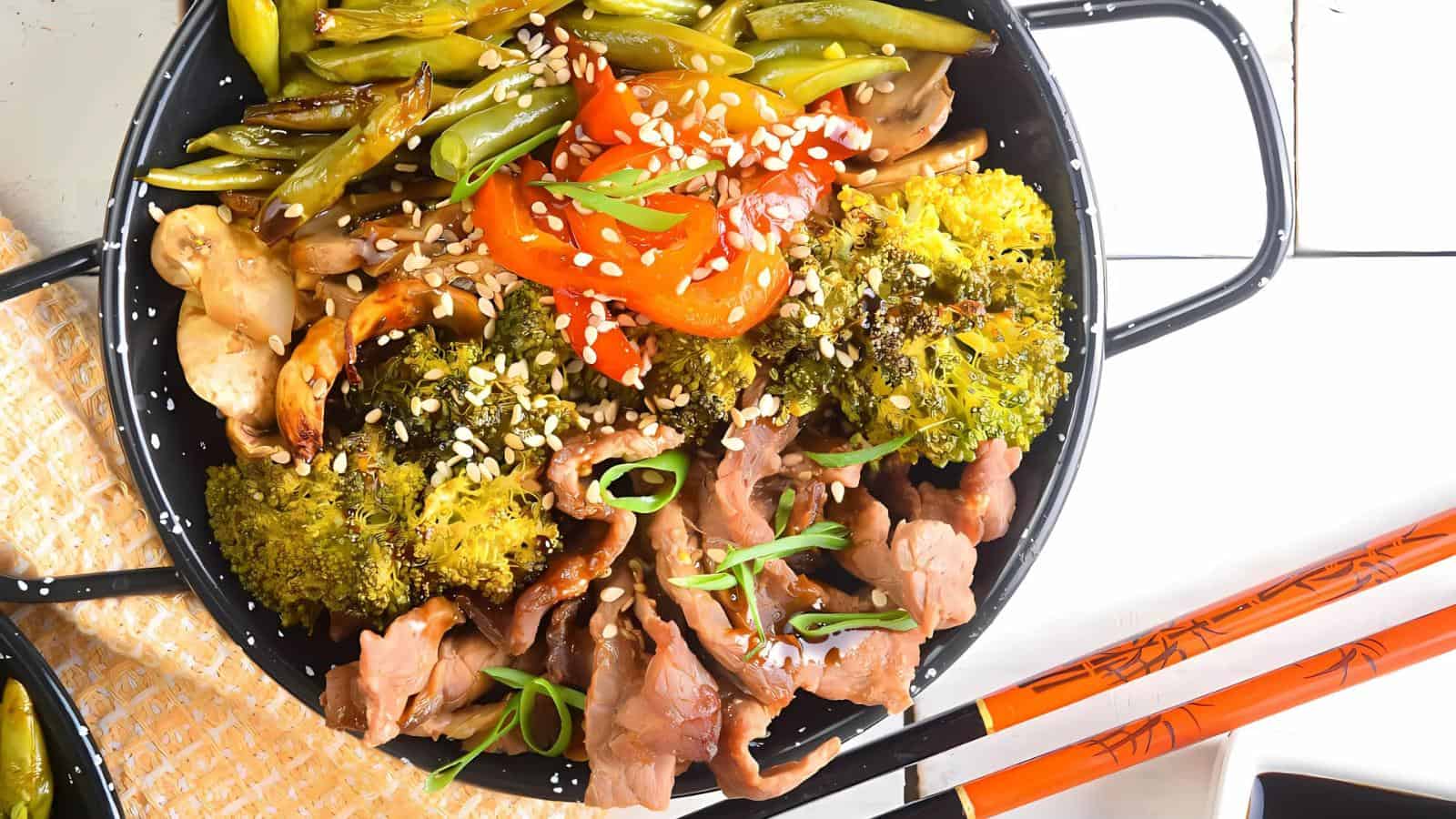 A bowl of stir-fried beef, broccoli, mushrooms, green beans, and red peppers, garnished with sesame seeds and green onions, with chopsticks on the side.