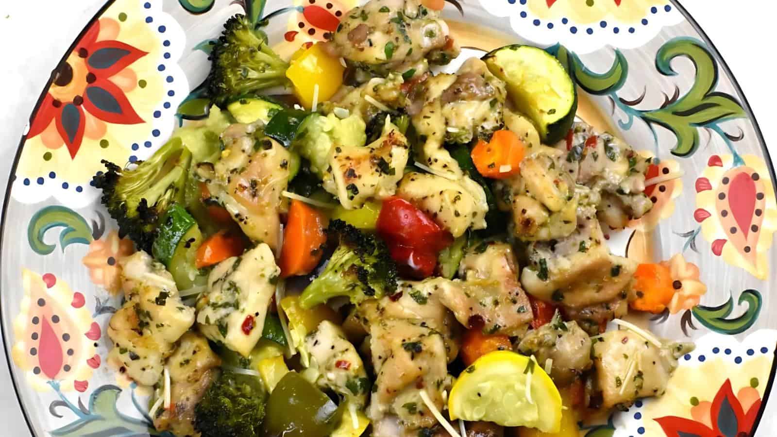 A colorful dish with cooked chicken pieces, broccoli, carrots, bell peppers, zucchini, and herbs on a decorative plate.