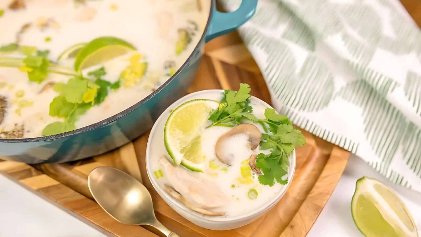 Bowl of creamy soup with lime slices, mushrooms, and cilantro on top, next to a pot and a spoon on a wooden tray.