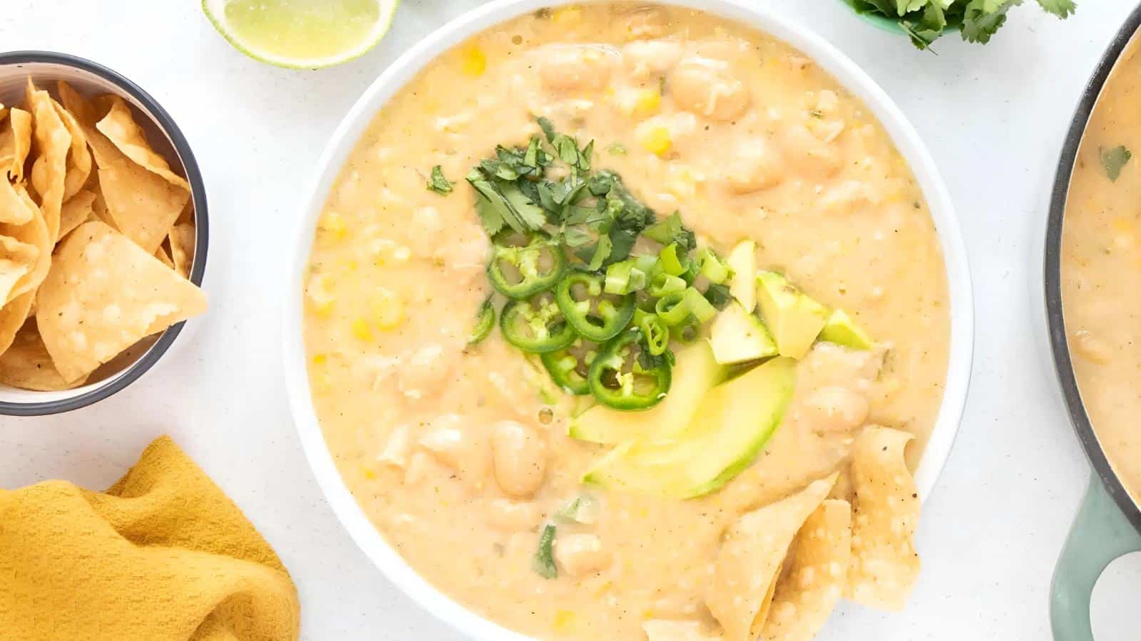 A bowl of creamy white bean chili topped with sliced avocado, jalapeños, and cilantro. Tortilla chips are partially dipped in the chili. A lime wedge and cilantro are nearby.
