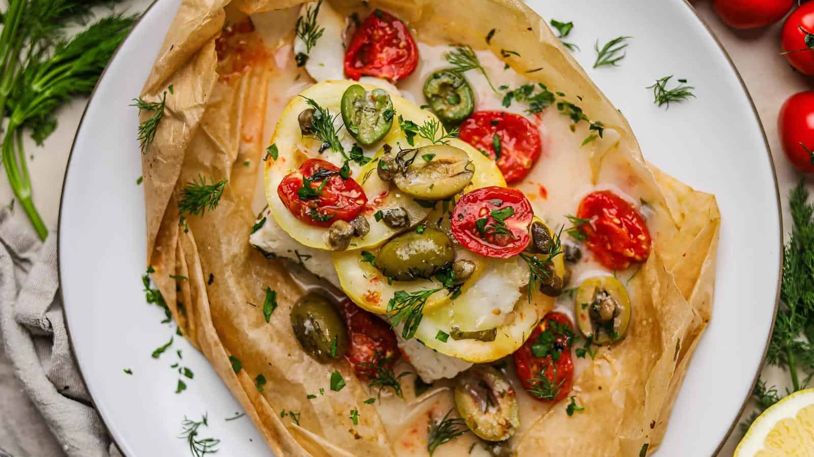 Baked fish with slices of lemon, green olives, cherry tomatoes, capers, and herbs on parchment paper in a white dish.