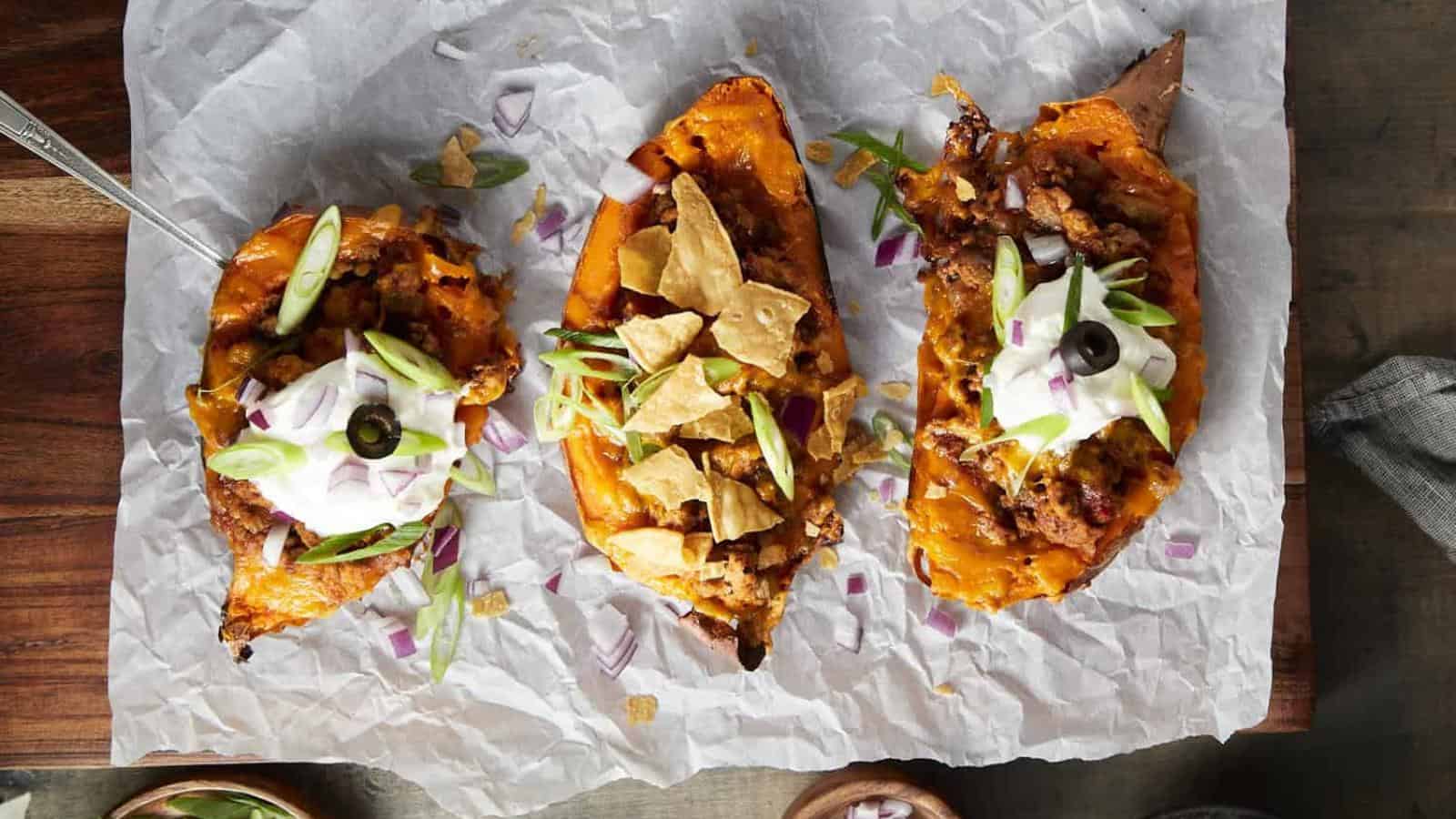 Three loaded sweet potato skins topped with sour cream, green onions, tortilla chips, and olives on parchment paper.